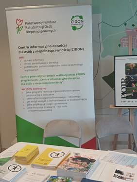 Pokaż zdjęcie: W tle baner z informacjami o CIDON w kolorach biało-zielonych, a na stole prze banerem ulotki informacyjne dotyczące programów PFRON.