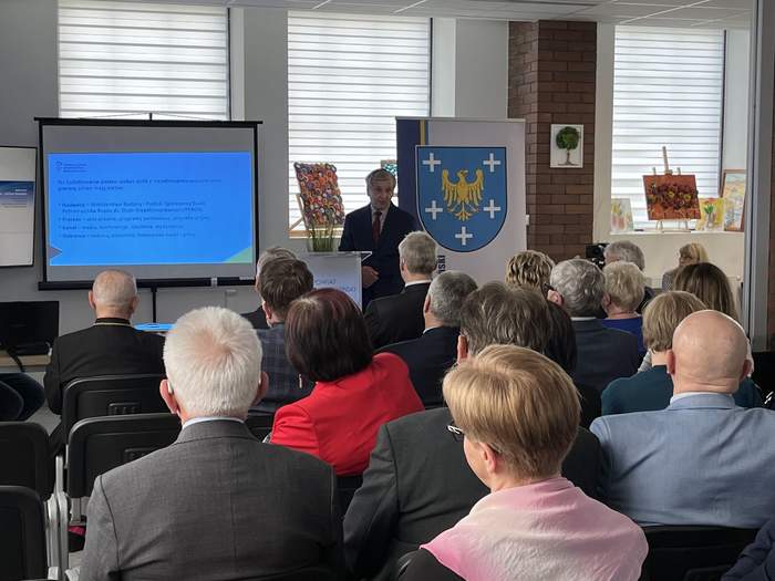 Pokaż zdjęcie: Fragment pomieszczenia na pierwszym planie osoby siedzą tyłem w rzędach w tle stoi mężczyzna, po lewej stronie stoi ekran z wyświetlaną prezentacją po prawej stoi baner z herbem powiatu