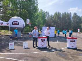 Pokaż zdjęcie: Dwóch mężczyzn stoi przy stolikach z logo wydarzenia sportowego "Start", jeden z nich mówi do mikrofonu. Za nimi widoczna jest dekoracja w formie dużej, nadmuchiwanej piłki z logotypami oraz tablica z logotypami sponsorów.