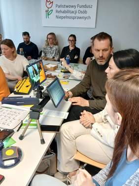 Pokaż zdjęcie: Grupa osób siedzących przy stole konferencyjnym.  Po prawej stronie trzy osoby testują powiększalnik zamocowany na statywie na stole. Nad powiększalnikiem znajduje się drugi ekran, również na ramieniu statywu, ustawiony pionowo, z kolorową zawartością na wyświetlaczu.