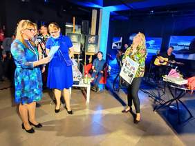 Pokaż zdjęcie: Na środku jubilatka - Monika Meleń, trzyma prezent od PFRON. Dookoła niej osoby, które składają jej życzenia