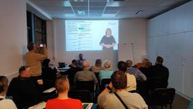 Pokaż zdjęcie: Grupa osób uczestniczy w warsztacie, siedząc w rzędach i słuchając prowadzącego, który znajduje się przed nimi. Na ekranie wyświetlany jest slajd z formularzem oraz nagranie z tłumaczką języka migowego.