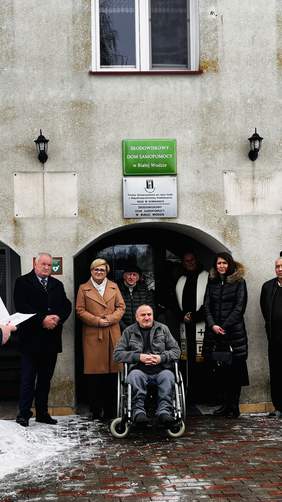 Pokaż zdjęcie: Uczestnicy wydarzenia stoją przed wejściem do Środowiskowego Domu Samopomocy w Białej Wodzie. W centrum znajduje się mężczyzna poruszający się na wózku inwalidzkim. Obok przedstawiciele instytucji, w tym Dyrektor PFRON Oddział Podlaski Edyta Kondzior oraz zaproszeni goście. 