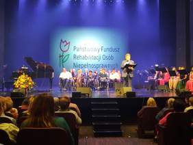 Pokaż zdjęcie: Na scenie w teatrze stoi Krzysztof Cwynar. Za nim znajdują się artyści ze Studia Integracji. Na widowni siedzi publiczność. W tle widać wielki napis: Państwowy Fundusz Rehabilitacji Osób Niepełnosprawnych. 