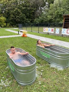 Pokaż zdjęcie: Na trawie stoją dwie balie z lodowatą wodą. Na tafli wody unoszą się kostki lodu. W jednej balii leży kobieta, a w drugiej mężczyzna. 