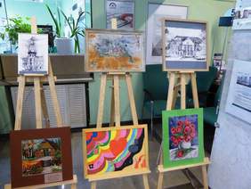 Pokaż zdjęcie: Na zdjęciu sześć prac stojących na sztalugach, przedstawiających grafiki architektury oraz kwiaty i serca z nutami. Prace stoją na korytarzu placówki.