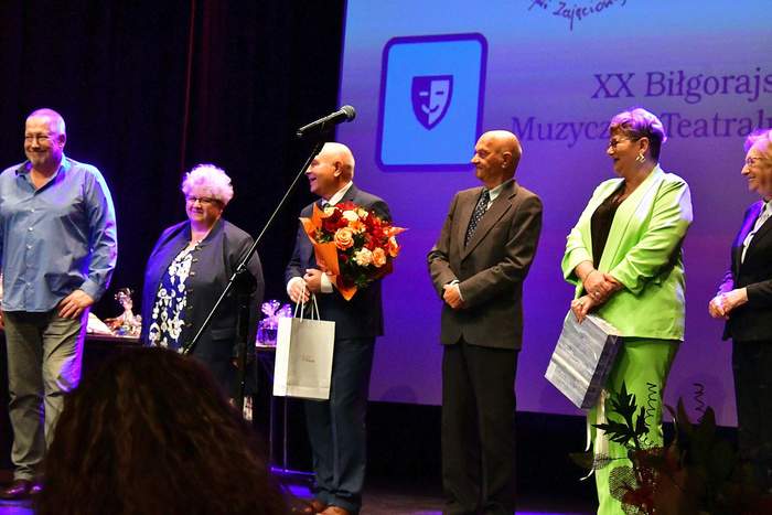 Pokaż zdjęcie: Na scenie stoi grupa sześciu osób elegancko ubranych, mężczyzna w garniturze z kwiatami składa życzenia jubilatowi.