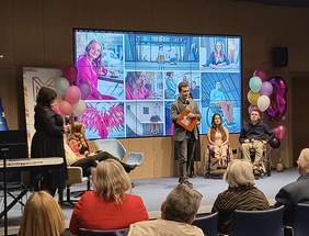 Pokaż zdjęcie: Prowadzący przemawia na scenie obok siedzących uczestników panelu, w tym osoby na wózkach inwalidzkich. Na ekranie za nimi wyświetlane są zdjęcia przedstawiające osoby związane z działalnością fundacji. Po bokach sceny dekoracje z balonów i elementy nawiązujące do fundacji MOŻESZ.