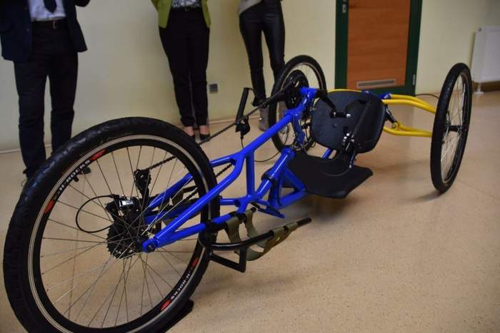 Pokaż zdjęcie: Rower trójkołowy niebiesko-żólty na tle sali i gości