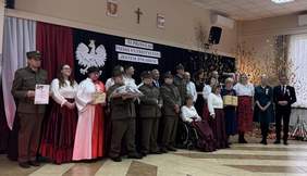 Pokaż zdjęcie: Grupa osób, część z nich w historycznych strojach wojskowych i ludowych, pozuje na scenie podczas XI Przeglądu Pieśni Patriotycznej „Jestem Polakiem” w Szumowie. W tle biało-czerwone dekoracje z orłem i napisami patriotycznymi. Na pierwszym planie widać osoby z dyplomami i upominkami.
