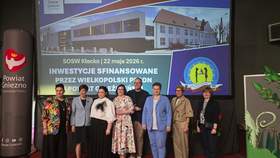 Grupa ośmiu osób stoi w jednym rzędzie na czerwonej scenie na tle dużego ekranu multimedialnego. Na slajdzie widoczna jest wizualizacja budynku oraz napisy: „SOSW Kłecko i  i data 22 maja 2026 roku, a poniżej napis: „Inwestycje sfinansowane przez wielkopolski PFRON”. Po lewej stronie sceny stoi pionowy baner Powiatu Gniezno z czerwonym herbem, a po prawej dekoracyjny panel z motywem zielonych liści.