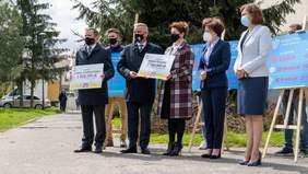 8 osób, 3 mężczyzn, 3 kobiety, z tyłu zarys jeszcze dwóch sylwetekujęcie lekko z boku, dwie trzymają tabice z napisanymi kwotami 1 828 299 oraz 2 700 000 