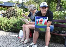 Pokaż zdjęcie: Uczestnicy warsztatów plastycznych. Dwie osoby siedzą na ławce i prezentują swoje obrazy