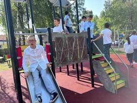 Pokaż zdjęcie: Na placu zabaw ze zjeżdżalni zjażdża chłopiec. Obok niego stoją inne dzieci. Pogoda jest słoneczna, w tle widać zieleń. 