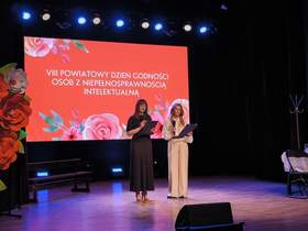 Dwie osoby stoją na scenie i czytają z kart, trzymając mikrofony. Za nimi duży ekran z napisem „VIII Powiatowy Dzień Godności Osób z Niepełnosprawnością Intelektualną” oraz dekoracją kwiatową i oświetleniem sceny.