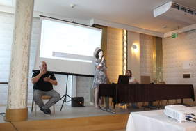 Pokaż zdjęcie: W sali konferencyjnym po lewej stronie siedzi na krześle tłumacz języka migowego, po prawej za stołem prezydialnym siedzi kobieta, druga stoi z mikrofonem, w tle po lewej na ekranie wyświetlana jest prezentacja