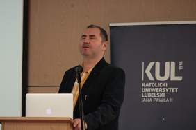 Pokaż zdjęcie: Przy mównicy stoi niewidomy mężczyzna ubrany w koszulę i marynarkę