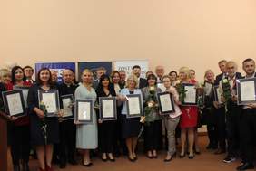 Pokaż zdjęcie: Na scenie stoją kobiet i mężczyźni, którzy trzymają w ręku białe róże oraz dyplom oprawiony w ramki