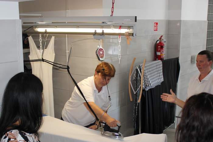 Pokaż zdjęcie: W pomieszczeniu znajduje się stanowisko do prasowania. Jedna kobieta trzyma w ręku żelazko i prasuje biały materiał, wokół wiszą ubrania. Trzy osoby stoją i się przyglądają.