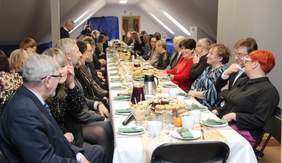 Pokaż zdjęcie: Przy długim stole siedzi wielu zaproszonych gości. Wszyscy są odświętnie ubrani. Na stole leży mnóstwo świątecznych potraw. 