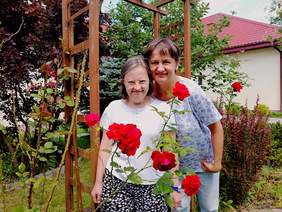 Pokaż zdjęcie: Dwie kobiety stoją w ogrodzie. Uśmiechają się. Przed paniami kwitnie czerwona róża. W tle widać budynek domu. 