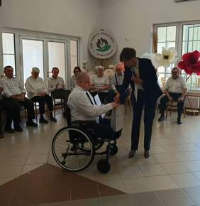 Pokaż zdjęcie: Uroczyste spotkanie w jasnej sali Domu Pomocy Społecznej w Baszkowie. Na pierwszym planie kobieta w ciemnym garniturze (Anna Skupień, dyrektor Oddziału Wielkopolskiego PFRON) pochyla się i podaje dłoń starszemu mężczyźnie na wózku. W tle grupa mieszkańców siedzących w półokręgu obserwuje scenę. Na ścianie widoczne logo placówki oraz duże dekoracyjne kwiaty w barwach narodowych.