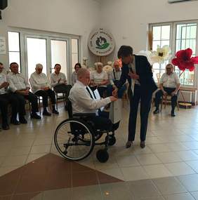 Pokaż zdjęcie: Uroczyste spotkanie w jasnej sali Domu Pomocy Społecznej w Baszkowie. Na pierwszym planie kobieta w ciemnym garniturze (Anna Skupień, dyrektor Oddziału Wielkopolskiego PFRON) pochyla się i podaje dłoń starszemu mężczyźnie na wózku. W tle grupa mieszkańców siedzących w półokręgu obserwuje scenę. Na ścianie widoczne logo placówki oraz duże dekoracyjne kwiaty w barwach narodowych.