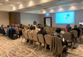 Sala konferencyjna, za mównicą stoi mężczyzna, przemawia, obok wyswietlona jest prezentacja, przed nim w kilku rzędach siedzi około 25 osób, widok z boku