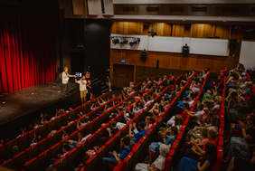 Pokaż zdjęcie: Na scenie stoi mężczyzna. Pod sceną stoi drugi mężczyzna, który trzyma w ręku laptopa. Za nim stoi  kobieta. Wszyscy są odwróceni w stronę widowni, która zapełniona jest publicznością