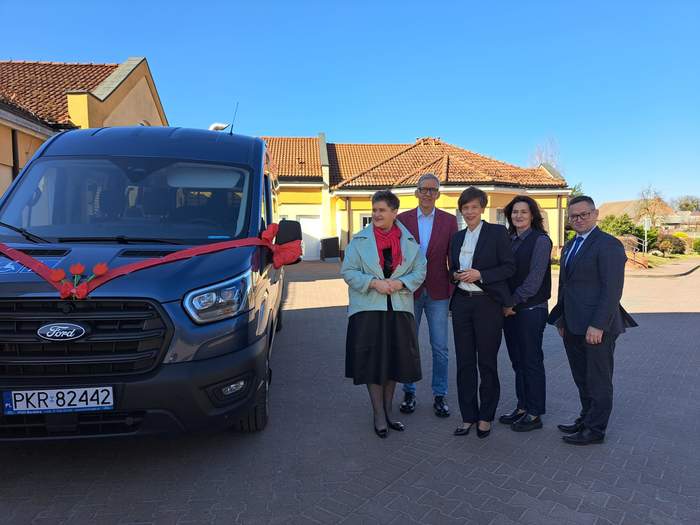 Pokaż zdjęcie: Grupa osób pozuje przy nowym, ośmioosobowym busie zaparkowanym przed budynkiem. Samochód zdobi czerwona wstążka na masce oraz czerwone tulipany. W tle dom z jasną elewacją i dużymi oknami.