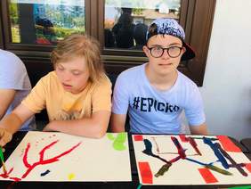 Pokaż zdjęcie: Dwóch chłopców - podopiecznych Stowarzyszenia "Tęcza" w takcie warsztatów plastycznych. Siedzą przy drewnianej ławie, malują farbami.