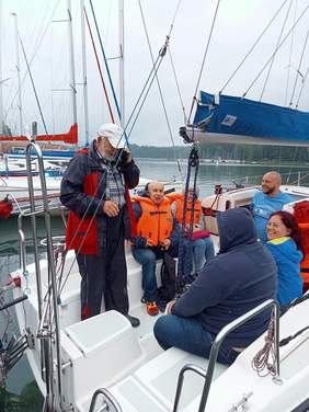 kilka osób na białym jachcie, męzczyzna z brodą stoi irozmawia przez telefon, mężczyzna i kobieta w pomarańczowych kapokach siedzą , na przeciwko nich 3 osoby bez kapoków, kobieta odwraca się i uśmiecha