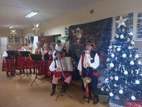 Pokaż zdjęcie: Występ zespołu ludowego w udekorowanej sali. Sześć kobiet w tradycyjnych strojach (czerwone spódnice w kwiaty, haftowane kamizelki) stoi przy pulpitach nutowych. Towarzyszą im muzycy: mężczyzna grający na kontrabasie, siedzący akordeonista oraz mężczyzna na pierwszym planie grający na diabelskich skrzypcach. Po prawej stoi ubrana choinka, a na ścianie wisi plakat ze Świętym Mikołajem.