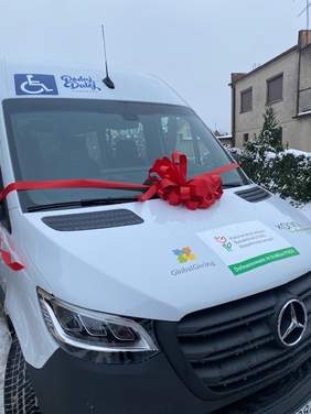 Pokaż zdjęcie: Biały bus z naklejkami informującymi, że pojazd dofinansowany został ze środków PFRON i jest dostosowany do przewozu osób na wózku. Dodatkowo na masce busa możemy dostrzec dwa logotypy: Miasta Konin oraz GlobalGiving. Pojazd został udekorowany także czerwoną kokardą.