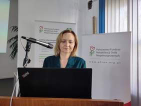 Pokaż zdjęcie: Za mównicą stoi kobieta, blondynka ubrana w zieloną sukienkę.