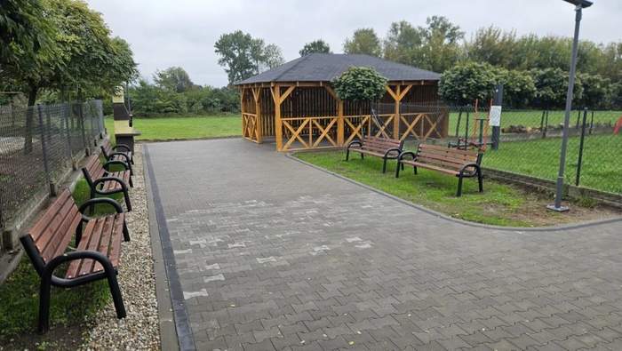 Pokaż zdjęcie: Szeroki chodnik prowadzący do grilla oraz altany grillowej. Po obu stronach chodnika umiejscowione zostały ławki, a w oddali znajdują się tereny zielone.