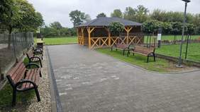 Pokaż zdjęcie: Szeroki chodnik prowadzący do grilla oraz altany grillowej. Po obu stronach chodnika umiejscowione zostały ławki, a w oddali znajdują się tereny zielone.