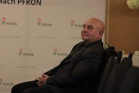 Pokaż zdjęcie: Na krześle w sali konferencyjnej siedzi starszy mężczyzna w garniturze, lekko się uśmiecha