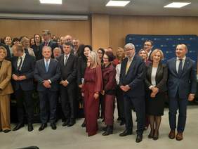 Pokaż zdjęcie: W sali stoją ludzie, którzy pozują wspólnie do zdjęcia. Wszyscy są elegancko ubrani. Mężczyźni mają na sobie garnitury, a kobiety garsonki i żakiety. 