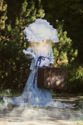 Pokaż zdjęcie: Fotografia przedstawiająca postać w jasnej sukni stojąca na leśnej ścieżce; zamiast głowy widoczna jest chmura, w dłoni postać trzyma małą kulę ziemską, a obok znajduję się duża walizka. 