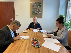 Pokaż zdjęcie: Przy drewnianym stole siedzi dwóch mężczyzn i kobieta. Na stole leżą dokumenty, pieczątki, cukierki i  filiżanka. Mężczyźni i podpisują dokumenty. Kobieta czyta dokument. Za nimi wisi obraz kolorowy w metalowej ramie.