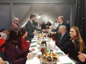 Pokaż zdjęcie: W sali jest ustawiony długi stół. Wokół niego siedzą ludzie. Niektórzy jedzą, a inni dzielą się opłatkiem lub składają sobie życzenia. 