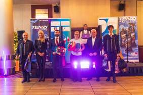 Pokaż zdjęcie: Grupa sześciu osób stoi na scenie podczas uroczystości wręczenia nagród. . Po lewej stronie stoją kobieta w czarnym stroju z bukietem kwiatów oraz młody mężczyzna w marynarce trzymający nagrodę i kwiaty. Po prawej stronie znajduje się trzech mężczyzn, jeden z nich (starszy, w okularach) trzyma bukiety kwiatów oraz torby upominkowe.