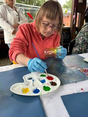 Pokaż zdjęcie: Kobieta w okularach i czerwonej polarowej bluzie maluje słoik farbami. Używa palety z różnokolorowymi farbami i trzyma pędzel w dłoni w niebieskiej rękawiczce.