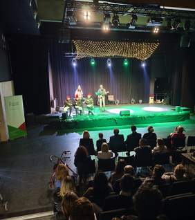 Pokaż zdjęcie: Szerokie ujęcie sali widowiskowej podczas występu. Na oświetlonej zielonym światłem scenie znajduje się pięciu wykonawców: czterech siedzi, a jeden stoi z gitarą. Trzech z nich ma na głowach czerwone czapki świętego Mikołaja. Nad sceną wiszą girlandy drobnych światełek przypominające gwieździste niebo. Na widowni, w przyciemnionej części sali, siedzi kilkanaście osób. Po lewej stronie widać zielony banner informacyjny PFRON.