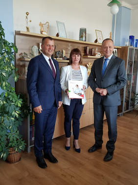 trzy osoby stoją na tle mebli w biurze, dwóch mężczyzn po bokach w środku kobieta, która trzyma dokumenty, wszyscy uśmiechają się