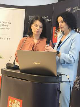 Pokaż zdjęcie: Przy mównicy stoją dwie prelegentki.