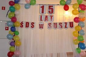 Pokaż zdjęcie: NA zdjęciu czerwony baner z napisem 15 lat ŚDS w Suszu, wokół niego kolorowe balony