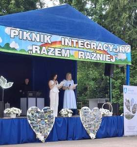 Scena plenerowa pod niebieskim namiotem; dwie osoby czytają z kartki, nad nimi baner „Piknik Integracyjny Razem Raźniej”, przed sceną dekoracje i kolaże zdjęć.
