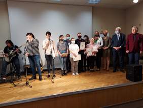Pokaż zdjęcie: scena na której śpiewają i grają na gitarze podopieczni WTZ Pokój w Łodzi oraz laureaci konkursu stoją z nagrodami obok dwie osoby wręczające nagrody i dyplomy 